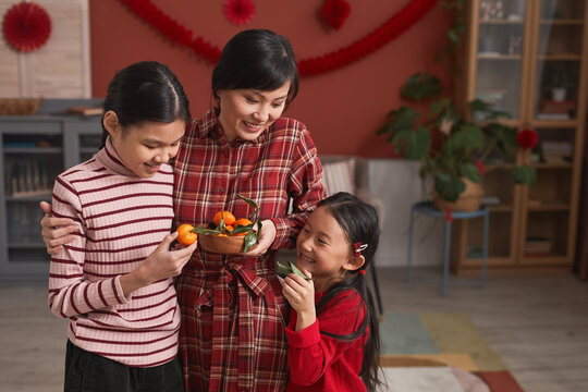 Family Portrait Of Chinese Woman And Two Lovely Daughters Standing Together In Living Room Taking Tangerines For Lunar New Year