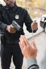 cropped view of african american policeman with handcuffs aiming by gun at blurred offender on foreground outdoors.