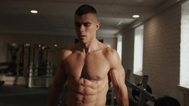 Young bodybuilder a with a naked torso training in the gym and does a standing exercise with dumbbells pumping biceps