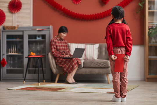 Horizontal long shot of little Asian girl standing in living room holding red envelope gift prepared for her mother behind her back