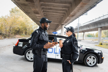 multicultural police officers with paper cups talking near patrol car on blurred background on urban street. © LIGHTFIELD STUDIOS