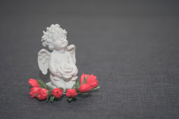Figurine of a white angel with a rose on a gray background. On the head there is an ornament in the form of a wreath of red flowers.