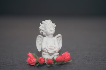 Figurine of a white angel with a rose on a gray background. On the head there is an ornament in the...