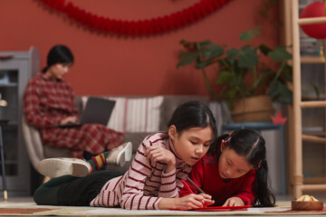 Portrait of two Chinese girls lying on floor in living room decorating handmade red envelopes with their drawings