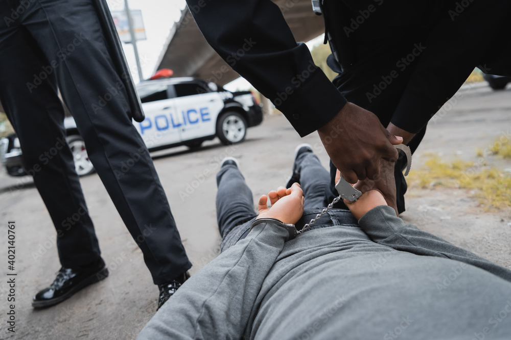 Wall mural cropped view of african american policeman handcuffing hooded offender lying on street on blurred background outdoors.