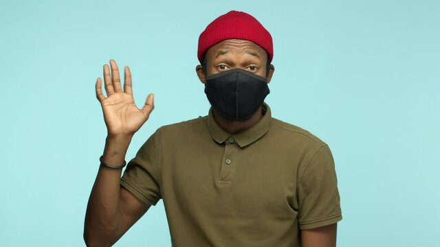 Coronavirus, pandemic and lockdown concept. Slow motion of friendly african american man wearing black face mask from covid, waving hand at camera and saying hello, blue background