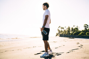 Male athlete in earphones contemplating sea standing on beach