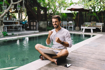 Young overjoyed businessman with modern laptop on poolside