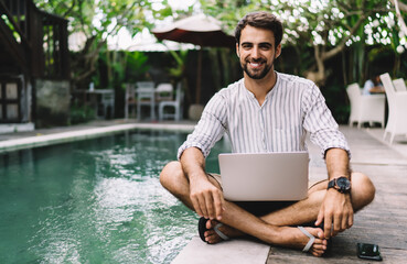 Cheerful casually dressed businessman with laptop near empty swimming pool
