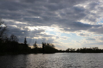 Naklejka premium The sun's rays make their way through the clouds in the sky on a spring evening on the shore of a reservoir