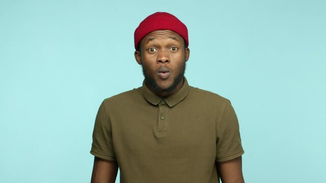 Slow motion of attractive african american man with beard, wearing red beanie and t-shirt, raising eyebrows and saying wow surprised, stare at impressive promotion, turquoise background