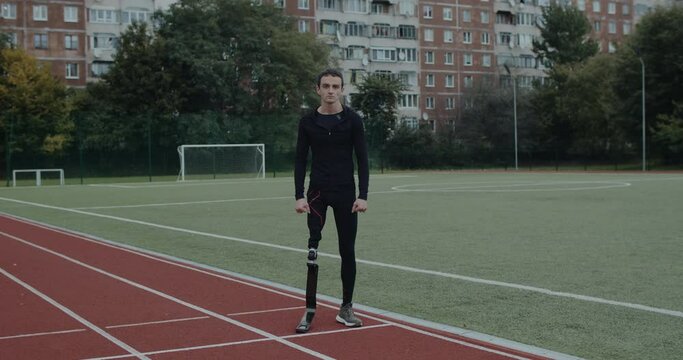 Handsome disabled man with prosthetic running blades turning and looking to camera. Amputee male runner standing at sports field outdoors. Concept of motivational sports footage.