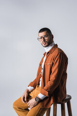 Fototapeta premium Stylish man in eyeglasses and terracotta jacket sitting on chair isolated on grey