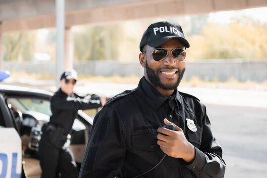 happy african american police officer looking at camera while holding radio set on blurred background outdoors.