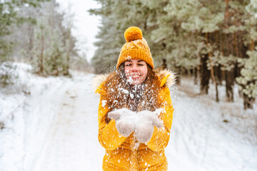 Beautiful young woman in yellow jacket and hat throws snow in snowy winter forest