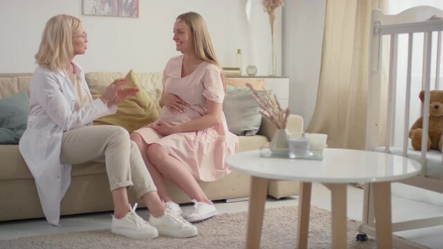Full shot of smiling female obstetrician-gynecologist in white scrubs and young expectant woman with big belly discussing pregnancy issues sitting at home in living room