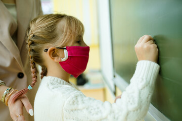 A girl with pigtails in a medical mask at the lesson writes in chalk on a blackboard