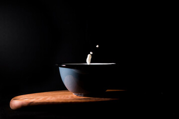 Drops of milk protrude from a bowl on black background. Copy space. Dark food concept.