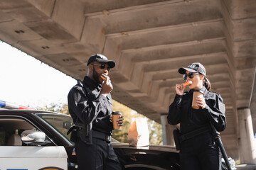 multicultural police officers with paper cups eating doughnuts near patrol car on blurred background on urban street. © LIGHTFIELD STUDIOS