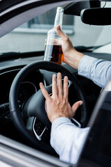 partial view of man beeping while driving car and holding bottle of whiskey, blurred foreground.