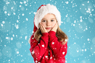 Cute child in Santa hat under snowfall on light blue background. Christmas celebration