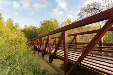 Side view of a red iron pedestrian bridge of a greenway surrounded by trees running over a river on...
