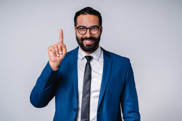 Smiling 25s middle eastern businessman in formal attire pointing at copy space isolated over grey background
