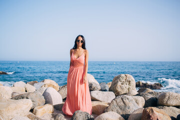 Full length portrait of beautiful female tourist in sundress and glasses for sun protection looking at camera while posing at coastline stones,charming woman in casual wear spending day for sunbathing