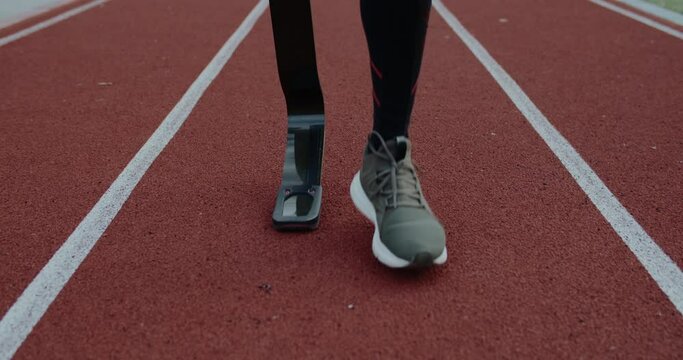 Crop view of unknown disabled male person with prosthetic running blades walking at sports field. Amputee male runner getting ready. Concept of motivational sports footage.