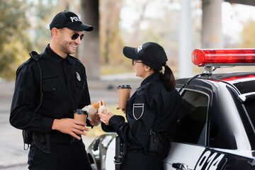 Smiling police officer holding takeaway coffee and burger near colleague during lunch near car outdoors.
