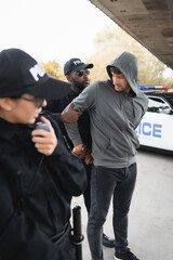african american policeman handcuffing hooded offender with blurred colleague on foreground outdoors.