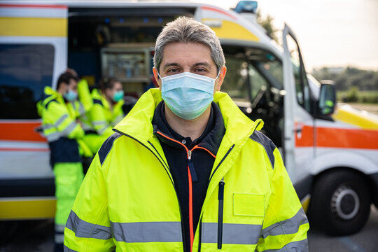 Portrait of friendly adult man with grey air at first aid in front of his colleagues in ambulance at break time at pandemic time from Coronavirus, Covid-19 He wearing face mask and uniform work