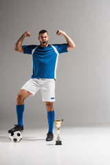 Excited sportsman showing yeah gesture near football and golden champions cup on grey background