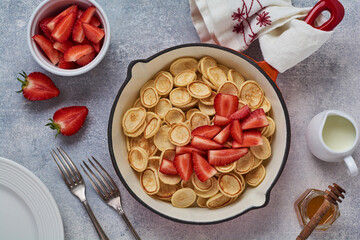 Mini white pancake cereal with strawberries in frying pan for breakfast on grey background . Trendy home breakfast with tiny pancakes. Top view.