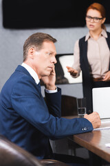 Frightened businessman sitting at workplace with blurred angry executive on background.