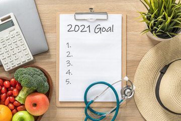 2021 goals on clipboard note pad for new year reminder list of yearly planner, health plan for work-life balance on desk with background of computer, calculator, travel hat and healthy food