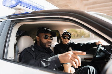 african american police officer pointing with finger near colleague in patrol car on blurred background. © LIGHTFIELD STUDIOS
