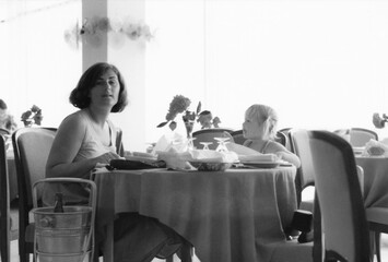 1977 vintage, seventies, retro monochrome image of a young mother and girl with blond hair sitting at a dining table in a restaurant of a hotel in Romania. © Studio F.