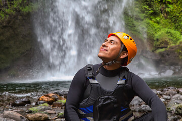 A closeup portrait of a cheerful man rock climber in safety harness sitting on a rock with a waterfall behind