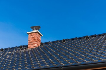 New chimney pipe in red bricks on roof in black shingles. On blue sky. Construction of a family house. Detail of the roof.