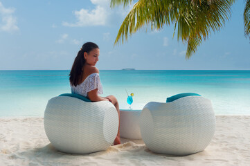Fototapeta na wymiar Brunette woman in white swimsuit drinking blue cocktail on the Maldive shore. White sand and turquoise ocean palm tree leaves. White rattan furniture.