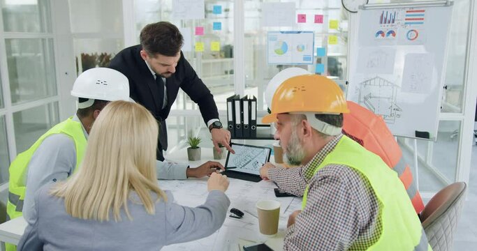 Good-looking confident purposeful diverse team of specialists of building industry holding a meeting and discussing details of joint building project when looking at tablet pc with depicted drawing
