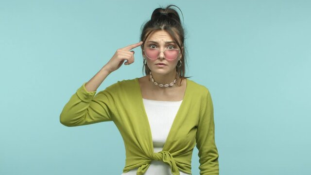 Slow motion of young brunette woman take-off sunglasses and stare at someone crazy acting stupid, pointing at head and camera, scolding idiot behaviour, standing over blue background