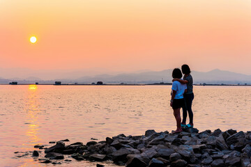 Mother and daughter two women standing watching the beautiful natural landscape on the waterfront, sun over the water in a bright orange sky at sunset on the lake, Krasiao Dam, Suphan Buri in Thailand