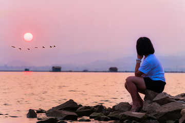 Asian teenage woman sit lonely alone at the waterfront looking at the beautiful nature landscape sun and flock of birds flying a row sunset at the lake background, Krasiao Dam, Suphan Buri, Thailand