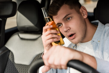 drunk man holding bottle of alcohol near head while driving car on blurred foreground.