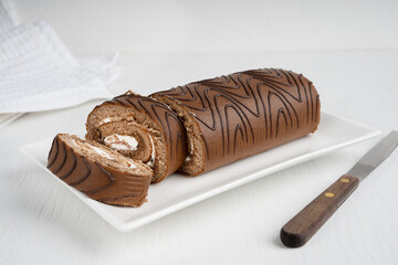 Chocolate homemade brown sponge roll cake or roulade with white cream filling sliced on rectangular plate with kitchen knife on white wooden table with textile towel at winter holidays. Horizontal