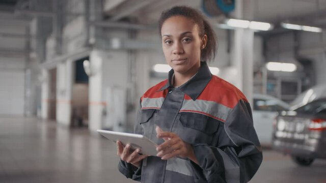 PAN portrait of black female auto mechanic in uniform using tablet while diagnosing problem in clients car in workshop, then looking at camera