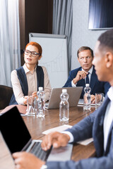 Redhead businesswoman talking while sitting at workplace with multicultural colleagues on blurred foreground in meeting room.