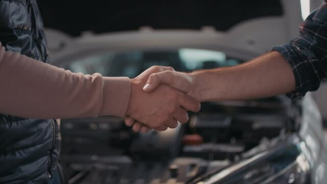 Close up slowmo of unrecognizable male client and male auto mechanic shaking hands. Broken down car with open hood and portable work light in background